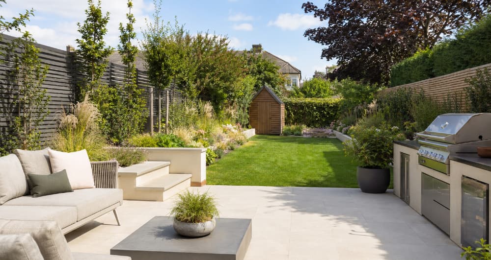 Contemporary landscaped garden with porcelain paving, outdoor kitchen, raised planting beds, horizontal slatted fencing and a timber garden shed positioned at the end of a neatly maintained lawn.