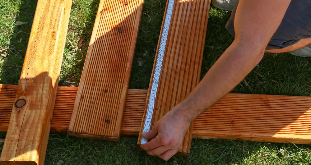 Person measuring wooden decking boards with a ruler during outdoor installation.