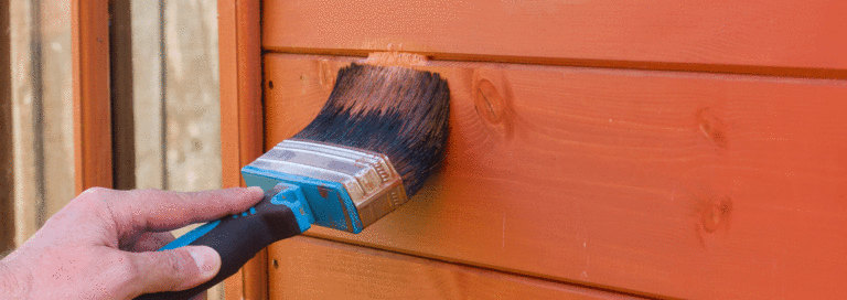 A close up of a hand holding a paintbrush to repaint a garden shed.